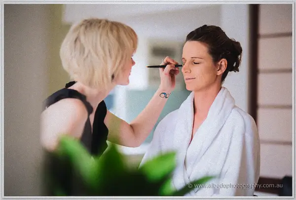 Bride in a white robe has her make-up finished by an artist beside a bright window before the ceremony.