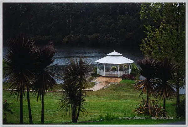 White rotunda beside a rain-flecked lake framed by grass trees on the Silkwood grounds.
