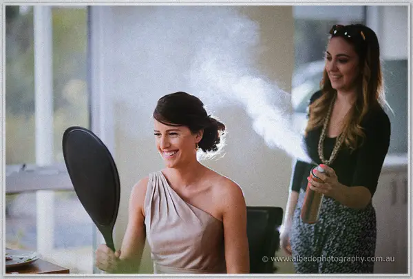 Laughing bride checks a hand mirror as hairspray mist drifts around her during morning preparations.
