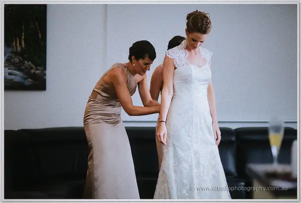 Bridesmaid in taupe smooths the lace skirt as the bride looks down at her gown beside a black sofa.