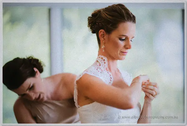 Bridesmaid leans in behind the bride as she checks a bracelet, lace-collared gown lit by window light.