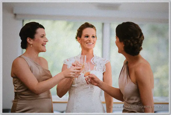 Bride and her two bridesmaids in gold clink champagne glasses and laugh by a bright window.