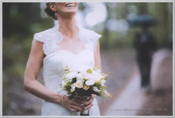 Bride laughs on a forest track clutching her bouquet as an umbrella holder waits behind in the drizzle.