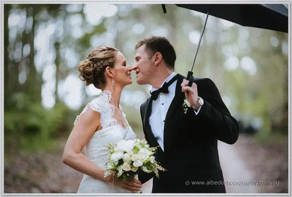 Groom under a black umbrella nuzzles his laughing bride's nose on a tree-lined forest path.