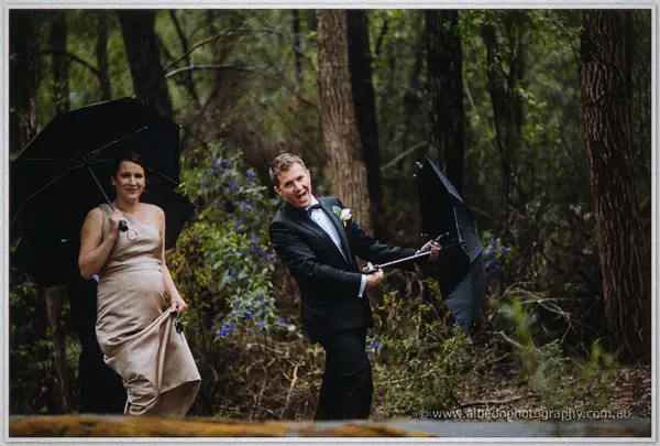 Groom wrestles an inside-out umbrella while a laughing bridesmaid ducks under hers in wet bushland.