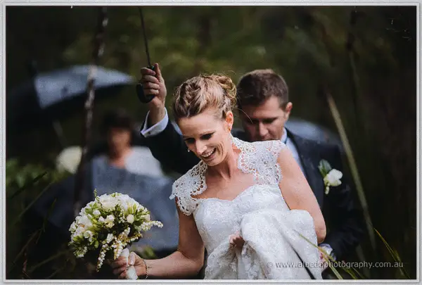 Beaming bride lifts her lace hem over wet ground as her groom shelters her beneath an umbrella.