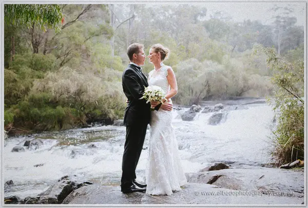 Bride and groom stand close on a granite rock beside a swollen, rain-swollen river rapid.