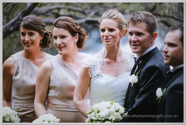 Bridal party in champagne gowns and dark suits gathers by the riverside, white bouquets in hand.