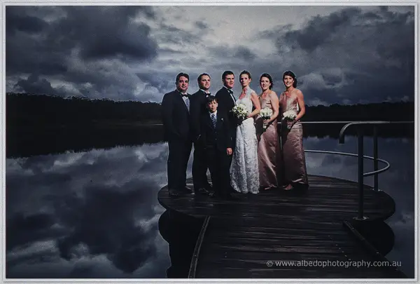 Bridal party and page boy line a wooden jetty under a brooding storm sky mirrored in still water.