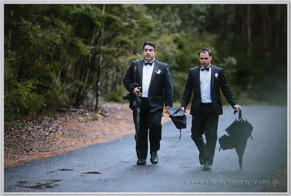 Two groomsmen in dinner suits stride down a wet forest road, umbrellas furled after the rain.