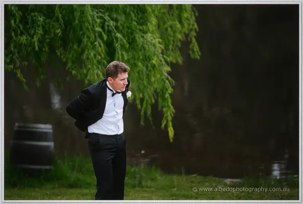 Groom adjusts his jacket under a willow beside the lake, wine barrel at his side.