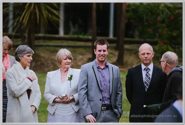 Wedding guests in coats and corsages chat on the lawn while light rain keeps falling.