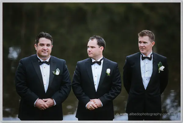 Groom and groomsmen in black bow ties and rose buttonholes wait solemnly by the lake.