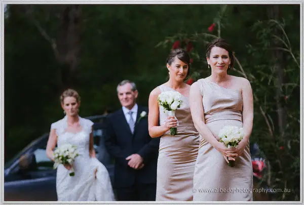 Bridesmaids in champagne satin lead the bride down the path toward the lakeside ceremony.