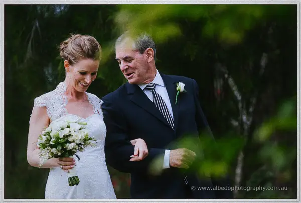 Smiling bride walks arm in arm with her father through soft green garden light.