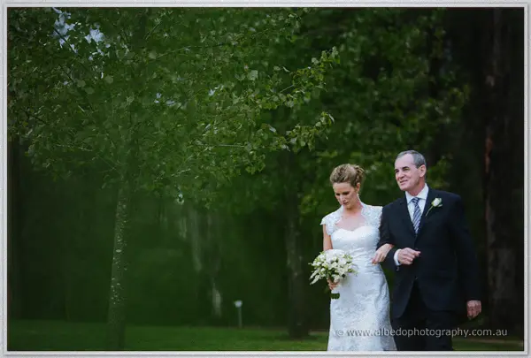 Bride on her father's arm crosses a rain-darkened lawn beneath fresh green canopy.