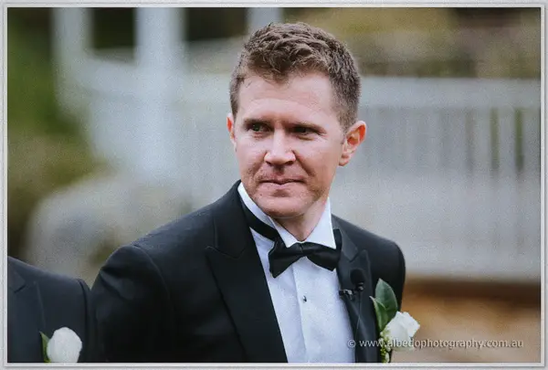 Teary groom in a bow tie fights back emotion as he waits at the ceremony by the water.