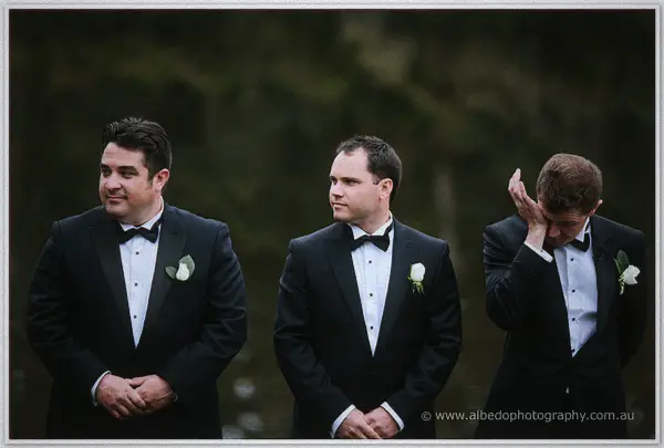 Groomsmen in tuxedos share the emotion, one covering his face as the bride approaches.