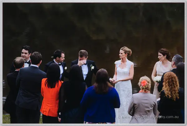 Guests gather waterside as the emotional groom covers his face beside his calm bride.