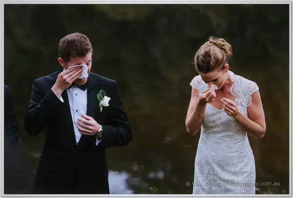 Bride and groom both dab their eyes with tissues during their lakeside wedding vows.