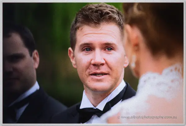 Wide-eyed groom looks up at his bride mid-vows, guests blurred in the forest backdrop.