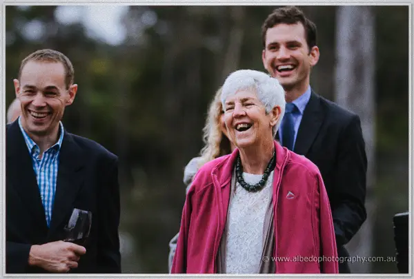 Wedding guests laugh openly during the ceremony, wine glass in hand under grey skies.