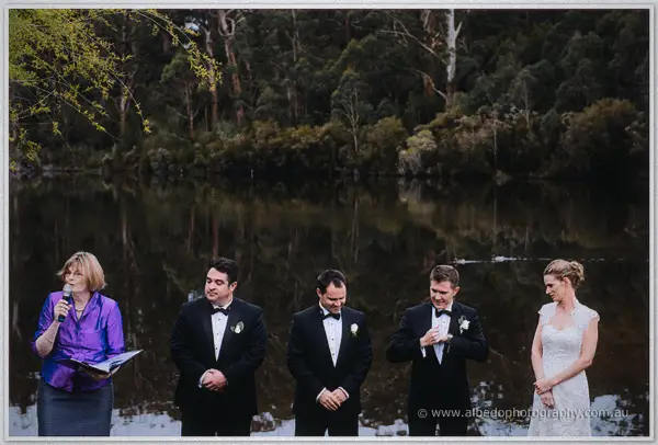Celebrant in purple leads the lakeside ceremony as tuxedoed groomsmen and the bride listen.