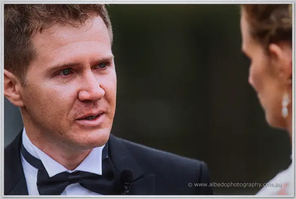 Groom in a bow tie speaks his vows, the bride's soft profile at the frame's edge.