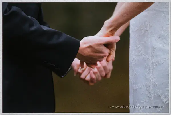 Close-up of the couple's clasped hands, his dark suit sleeve against her beaded lace gown.