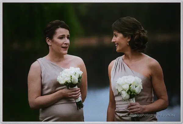 Two bridesmaids in champagne one-shoulder gowns laugh with white bouquets by the winery lake.