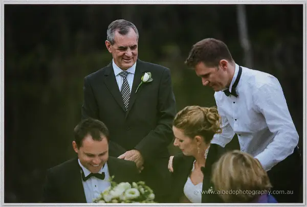 Beaming father figure in a suit with rose boutonniere greets the seated couple at the garden ceremony.