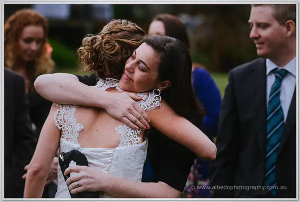 Bride in a lace-backed gown embracing a delighted guest among the winery garden crowd.