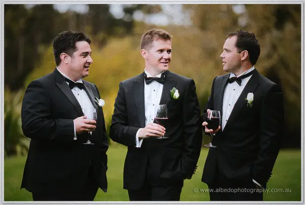 Groom and groomsmen in black tuxedos laughing over glasses of red wine in the garden.