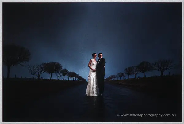 Bride and groom standing close on a rain-slicked tree-lined road under a stormy dusk.