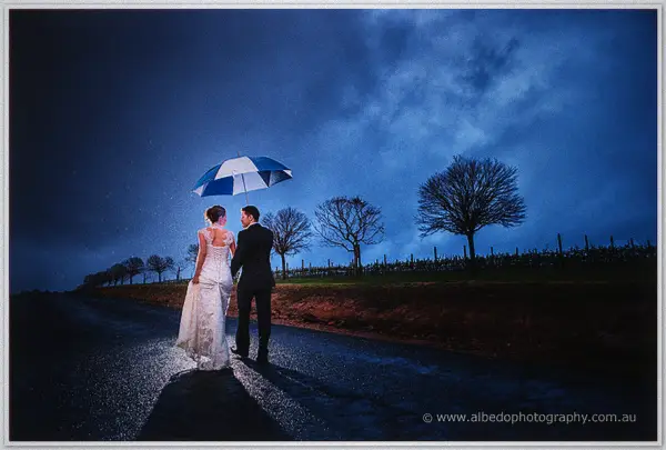 Bride and groom walk a wet vineyard road beneath a blue umbrella under a moody dusk sky