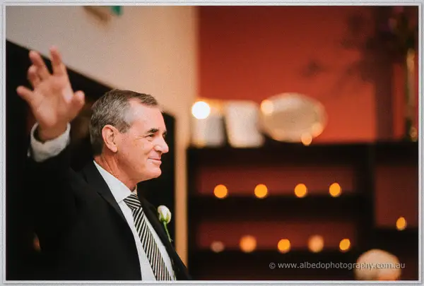 Father of the groom waving mid-toast, candles glowing on the shelves behind him.