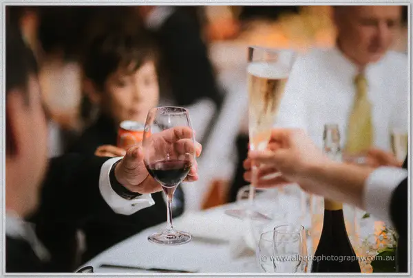 Guests' hands clinking red wine and champagne glasses over the candlelit reception table.