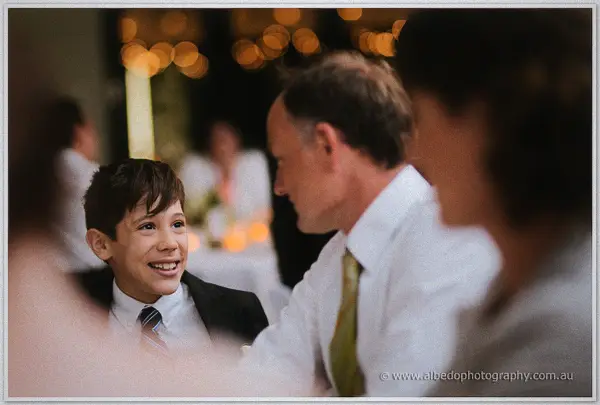 Young page boy in a suit grinning up at his father at the candlelit reception table.