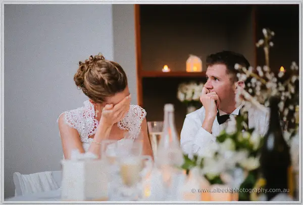 Bride covering her face in laughter as the groom listens wide-eyed to a toast.