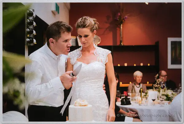 Groom pulling a playful face while starting the cake cut as wedding guests watch on.