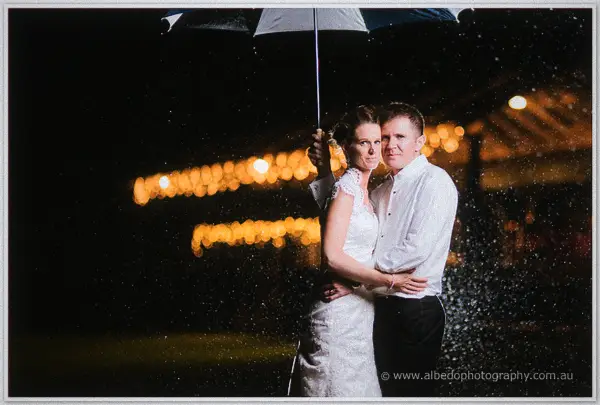 Newlyweds sheltering under an umbrella in night rain, festoon lights glowing behind them.