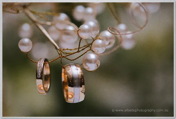 Two gold wedding rings hanging from pearl-studded branches against green bokeh.