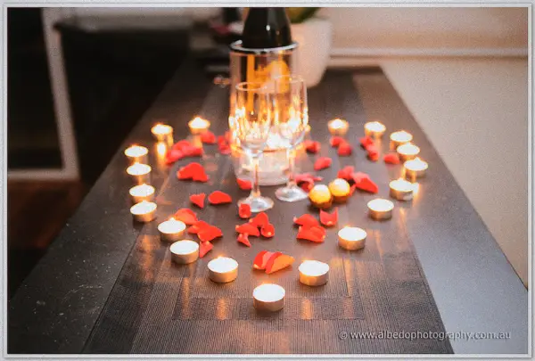 Tealights and rose petals circling two champagne flutes on a dark timber table.