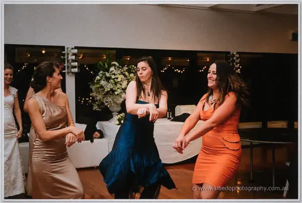 Three female guests in bronze, teal and orange dresses dancing in a row at the reception.