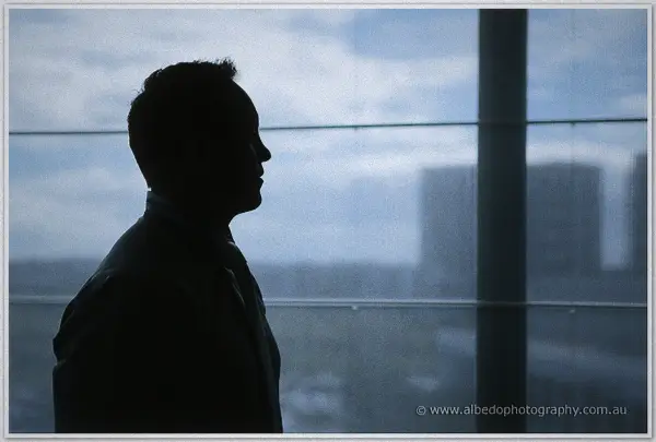 Nathan gazes out a floor-to-ceiling window, city towers soft in haze behind his morning silhouette