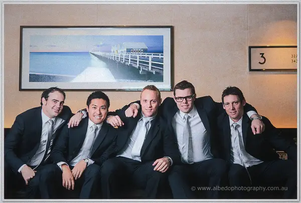 Five groomsmen in charcoal suits squeeze onto a lounge beneath a framed jetty print, grinning widely