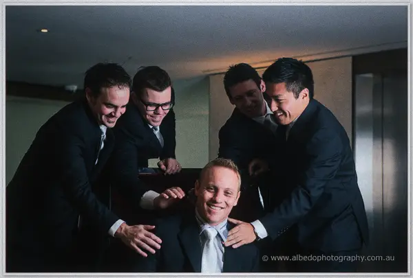 Four groomsmen in dark suits pile hands on Nathan's shoulders as they all laugh in the dim hotel room