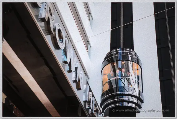 Nathan and his groomsmen ride a glass lift up a city tower facade framed in white and dark mullions