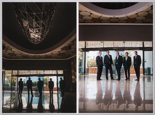 Groomsmen silhouetted beneath a sculptural lobby chandelier, then lined up on polished marble by glass doors