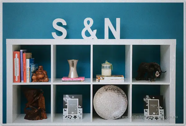A white shelf against a teal wall spells S and N in big letters above books, ornaments and frames